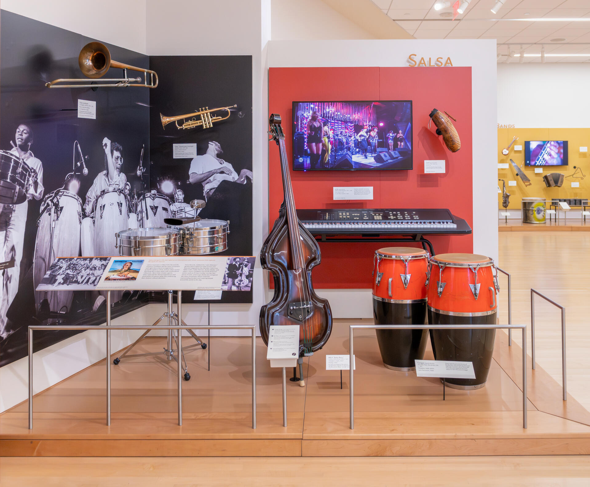 MIM's New Salsa Display Salsa exhibit from the Musical Instrument Museum showing congas, timbales, electric upright bass, trombone, trumpet, a digital keyboard, a guiro, video, and a graphic showing Ray Barretto and musicians.